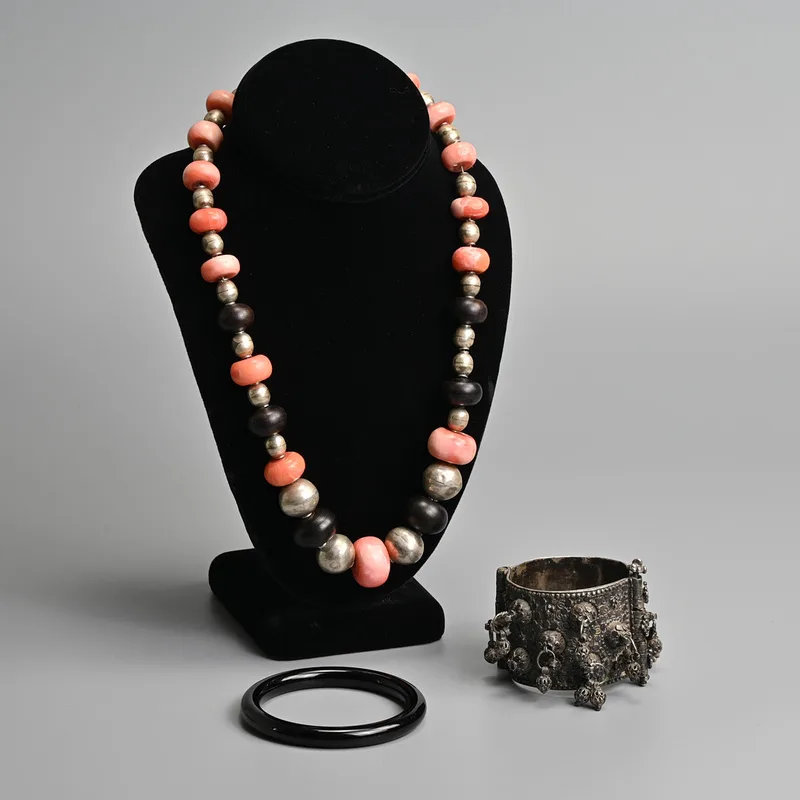 A Middle Eastern Coral and White Metal Beaded Necklace, Together with a Hinged White Metal Charmed Bracelet and a Black Hardstone Bangle